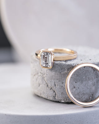 A Noam Carver emerald cut diamond engagement ring and gold wedding band are placed together on top of a grey piece of rounded stone and on white marble, fine jewelry by Noam Carver sold by Perrara.