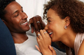 A man and woman laying closely next to each other and smiling at each other, both wearing beautiful wedding bands for her and him, while the woman also wears diamond earrings, a diamond necklace, and a diamond engagement ring by Tacori, sold at Perrara in